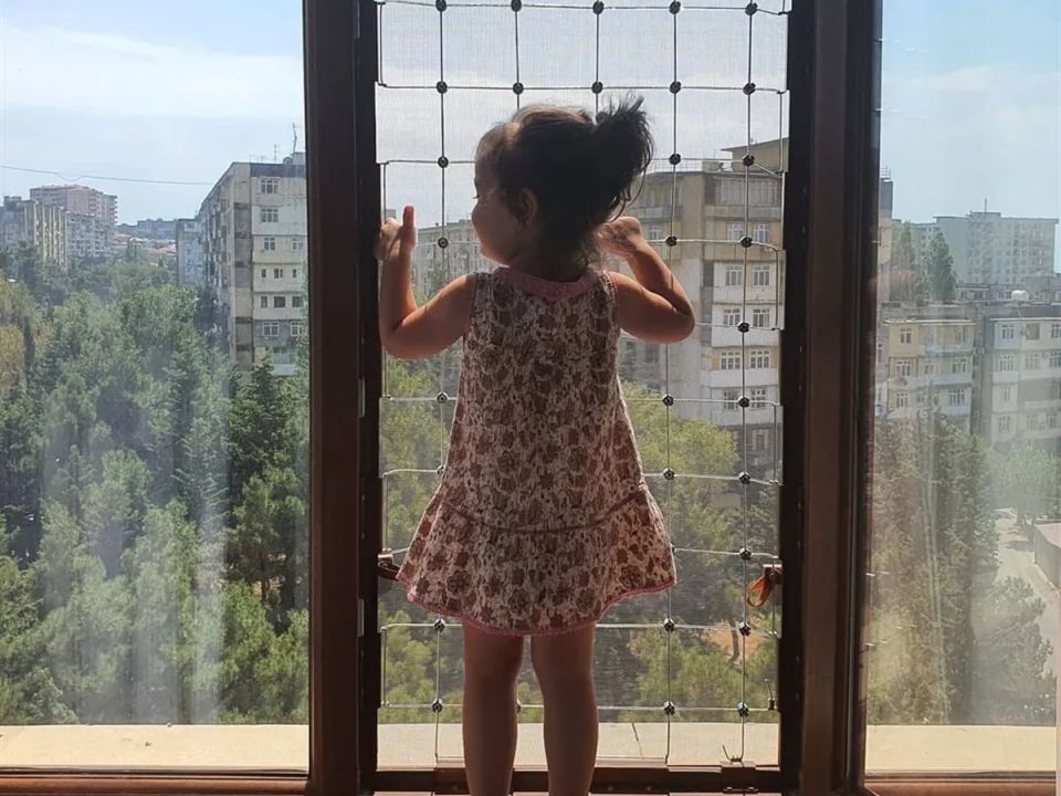 Açık pencereye gerilmiş geniş gözlü güvenlik filesinin önünde dışarıyı izleyen küçük çocuk