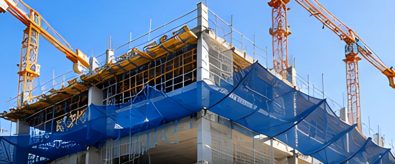 Vinçlerin yükselttiği binanın kat kenarlarında mavi inşaat güvenlik ağları