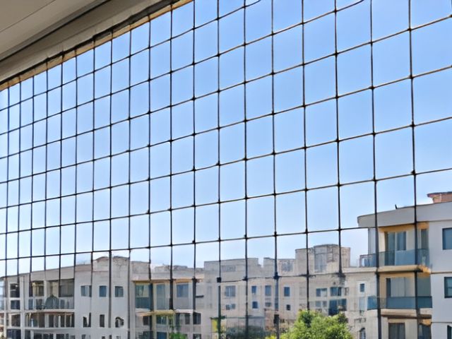 Geniş pencerenin önüne gerilmiş güvenlik filesi ve arkasında apartmanlar